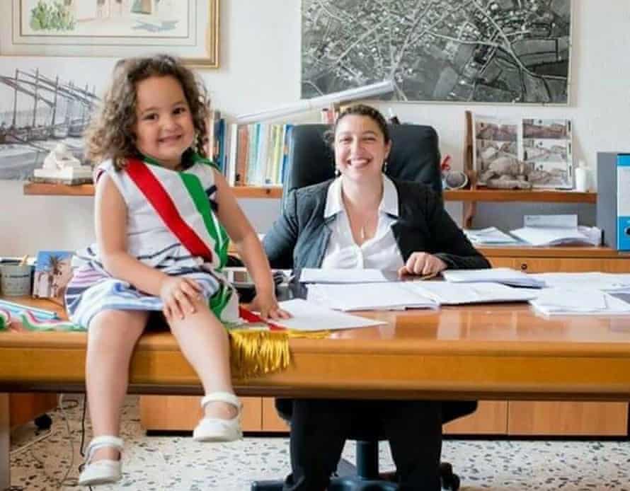 Debora Porrà at her desk with a smiling child sitting on it among her papers