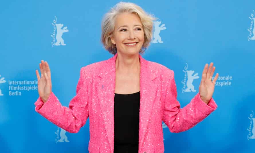 Emma Thompson in a pink blazer over a black pantsuit poses against a blue background with her hands out to either side.