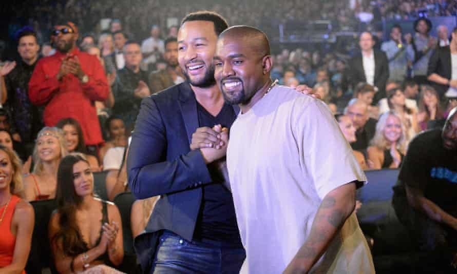 Two musicians, John Legend and Kanye West, shake hands during a performance in a crowded place.