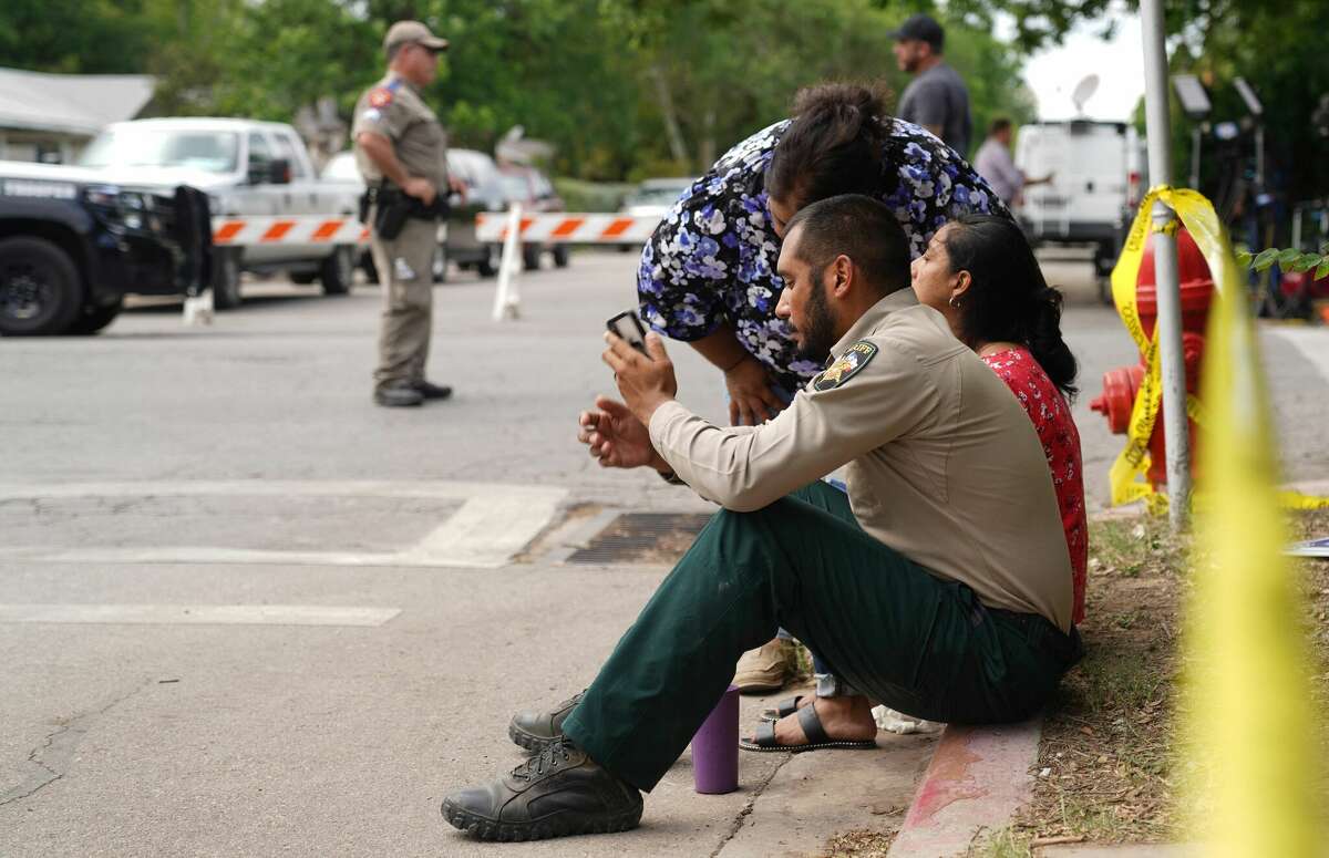 A sheriff checks his phone as he sits on the sidewalk with two women outside Robb Elementary School as state troopers monitor the area in Uvalde, Texas, May 24, 2022. - An 18-year-old gunman killed 14 children and a teacher at an elementary school in Texas on Tuesday, according to the state's governor, in the nation's deadliest school shooting in years. (Photo by allison dinner / AFP) (Photo by ALLISON DINNER/AFP via Getty Images)