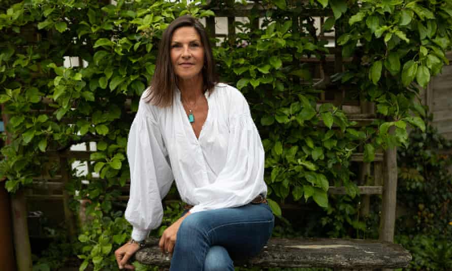 Lynn Wales today, sitting on a stone bench in front of a trellis covered in plants, with long dark hair and wearing a white shirt and jeans