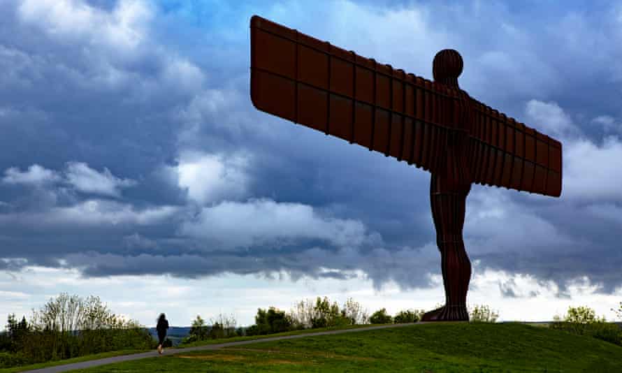 The Angel fo the North seen silhouetted from behind against a dramatic cloudy sky, with a lone jogger running past on the path beneath it