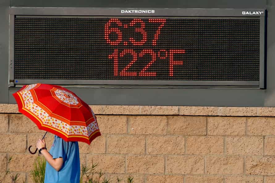 A person in a blue shirt carrying a red umbrella walks past a digital sign posting the temperature at 122F.