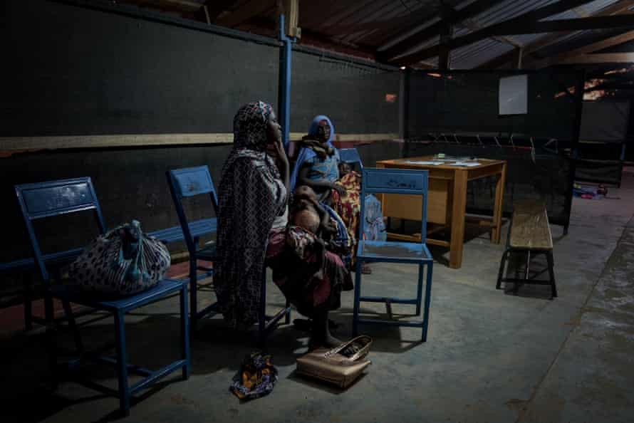 Two mothers sit with their small children on their laps in a dimly lit shed
