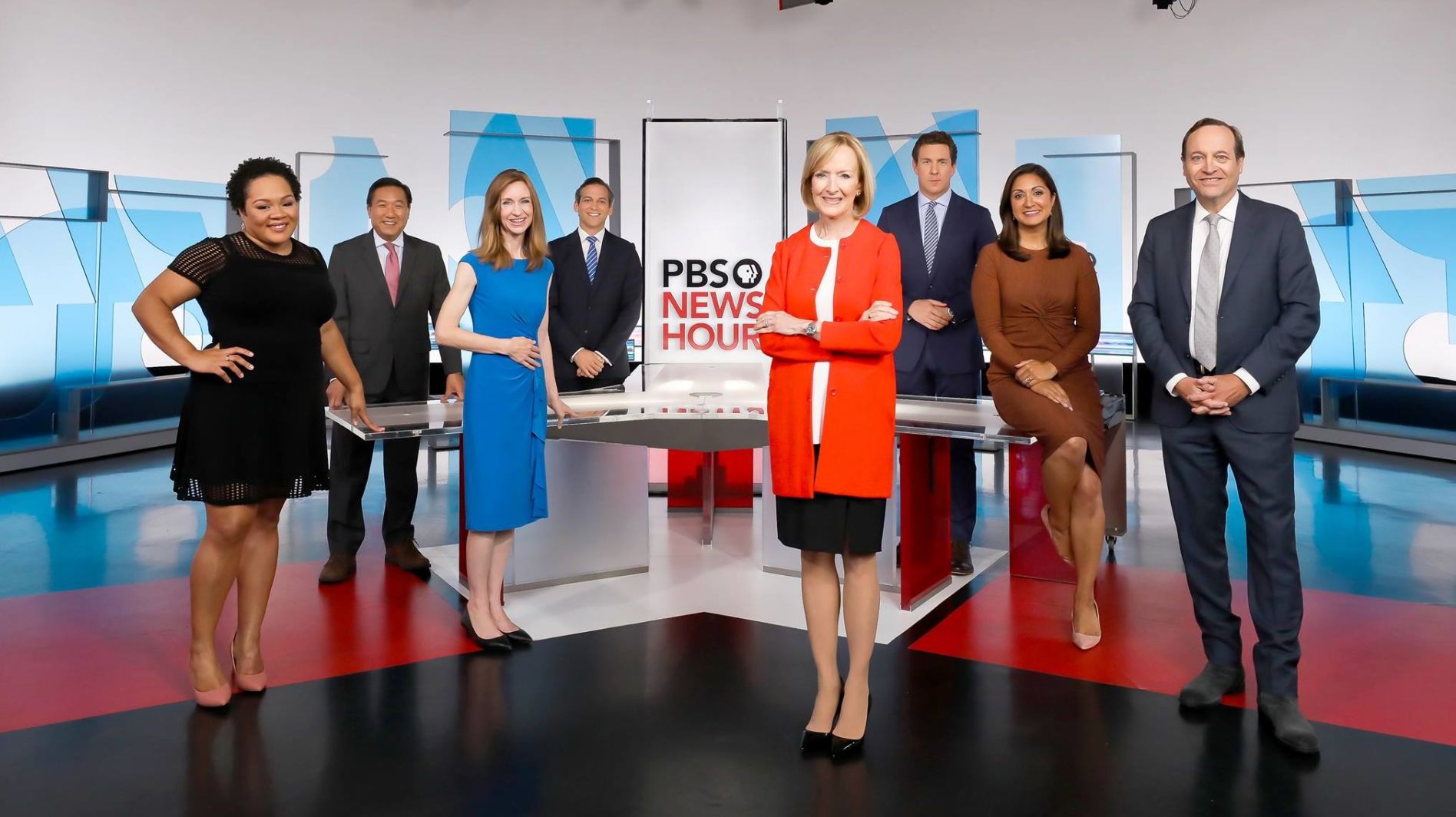 In a glossy TV studio, men and women in business pull spread out and pose for a publicity photo.  In the enter, an older woman in bright red jacket and black skirt folds her arms.